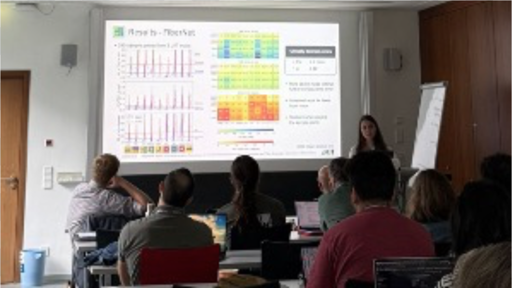 Person hält Präsentation mit Grafiken und Tabellen vor Publikum in einem Seminarraum.